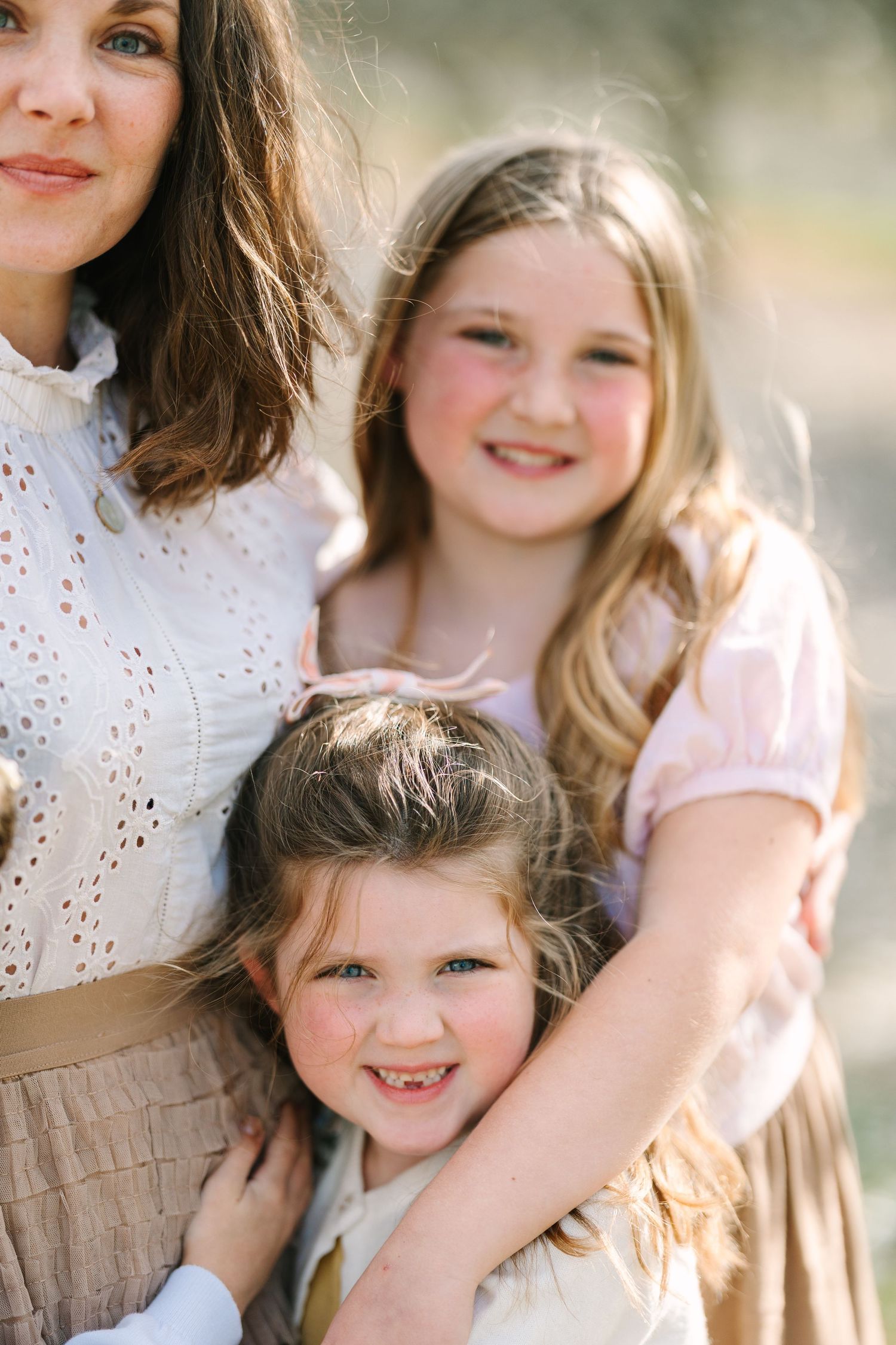 A mother and two young daughters smiling together outdoors in soft natural light, wearing pastel dresses.