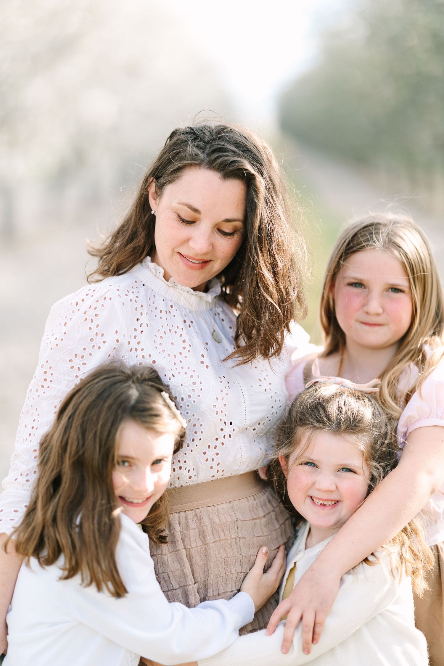 A mother smiles warmly while three young daughters embrace her outdoors in soft natural light.