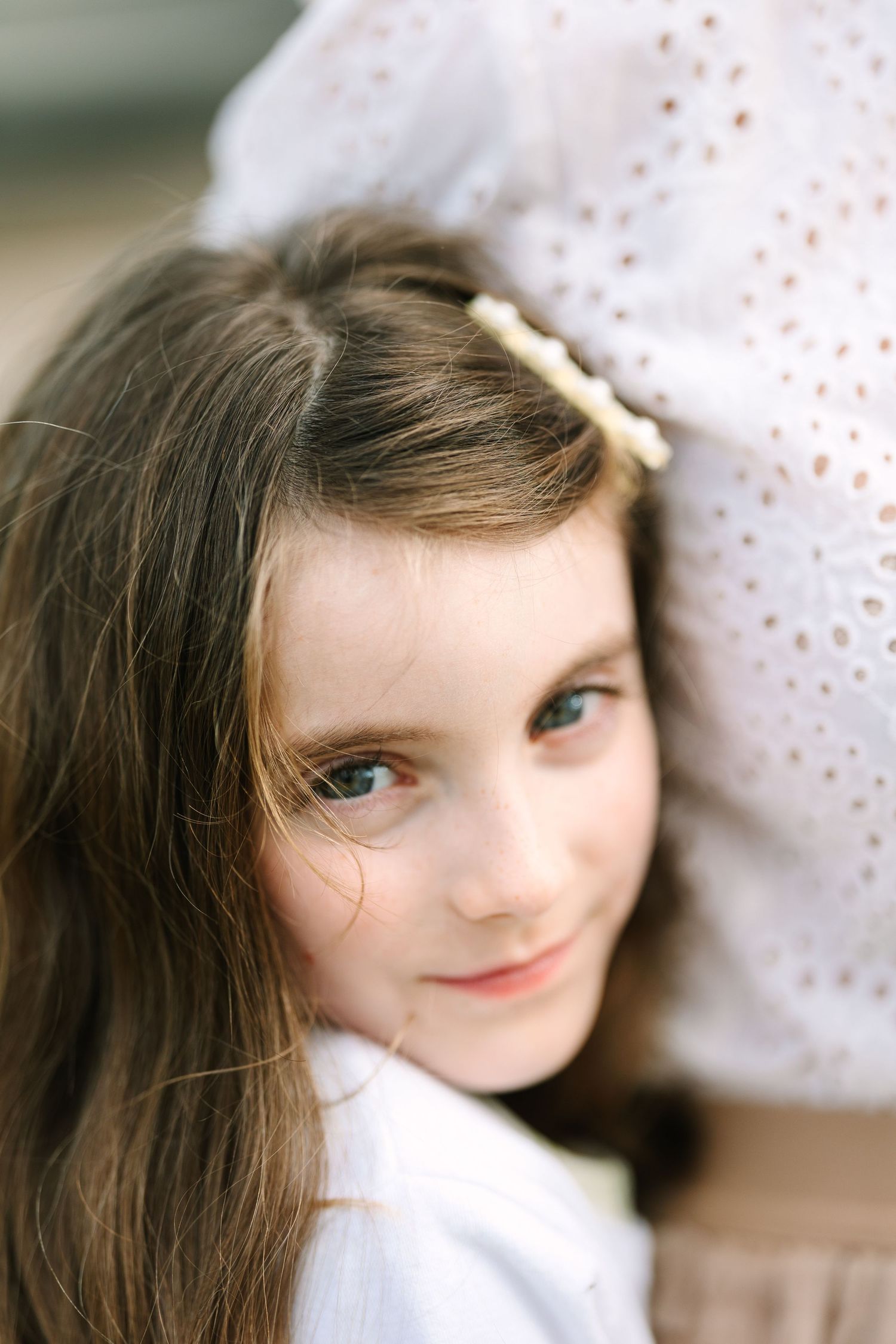 Young girl with long brown hair and a pearl hair clip smiles softly, wearing a white top outdoors.