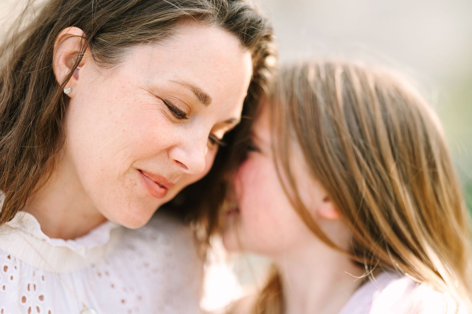 A mother and daughter share a tender close-up moment, touching foreheads gently outdoors in soft natural light.