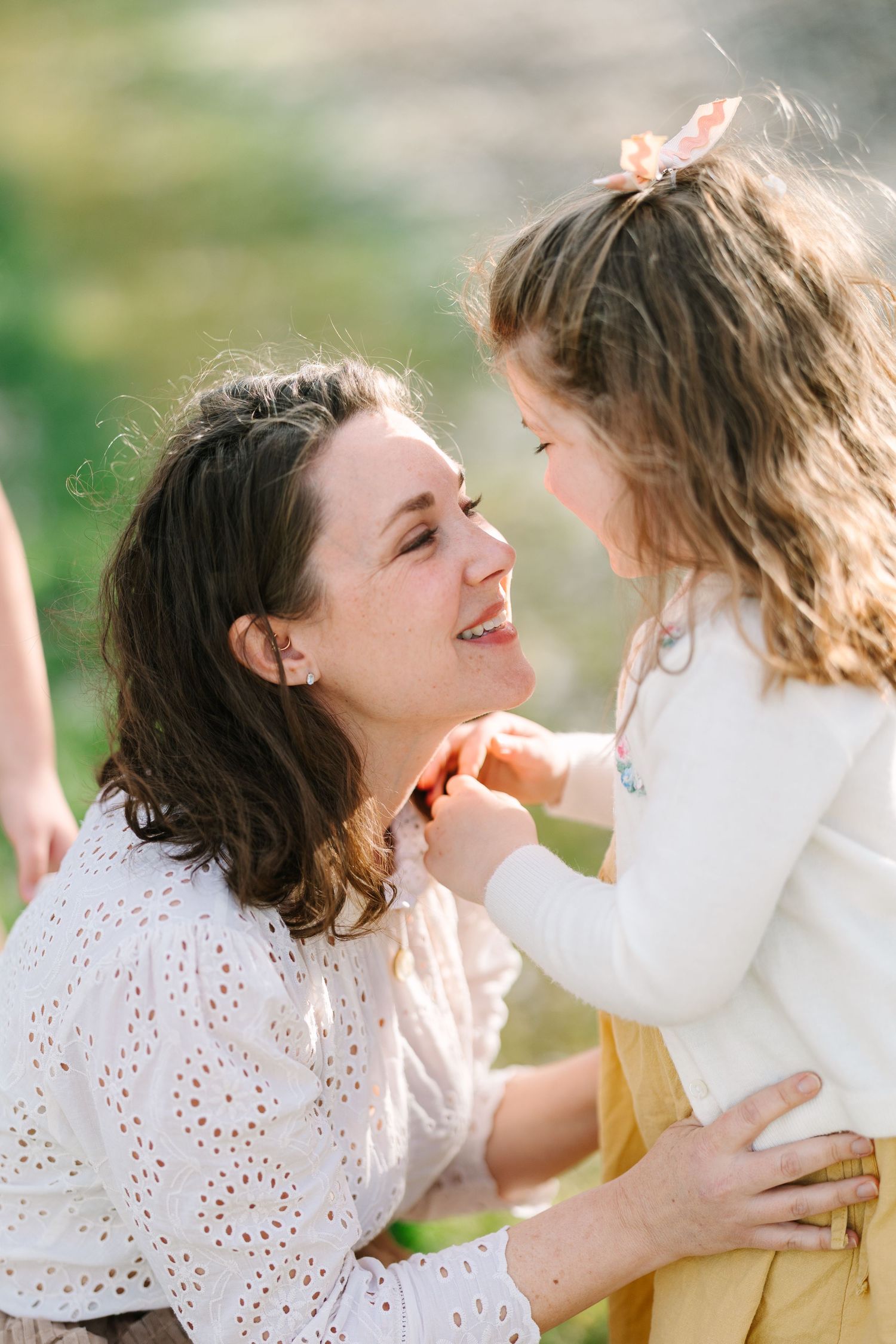 A mother and young daughter share a tender moment outdoors, smiling at each other in warm sunlight.