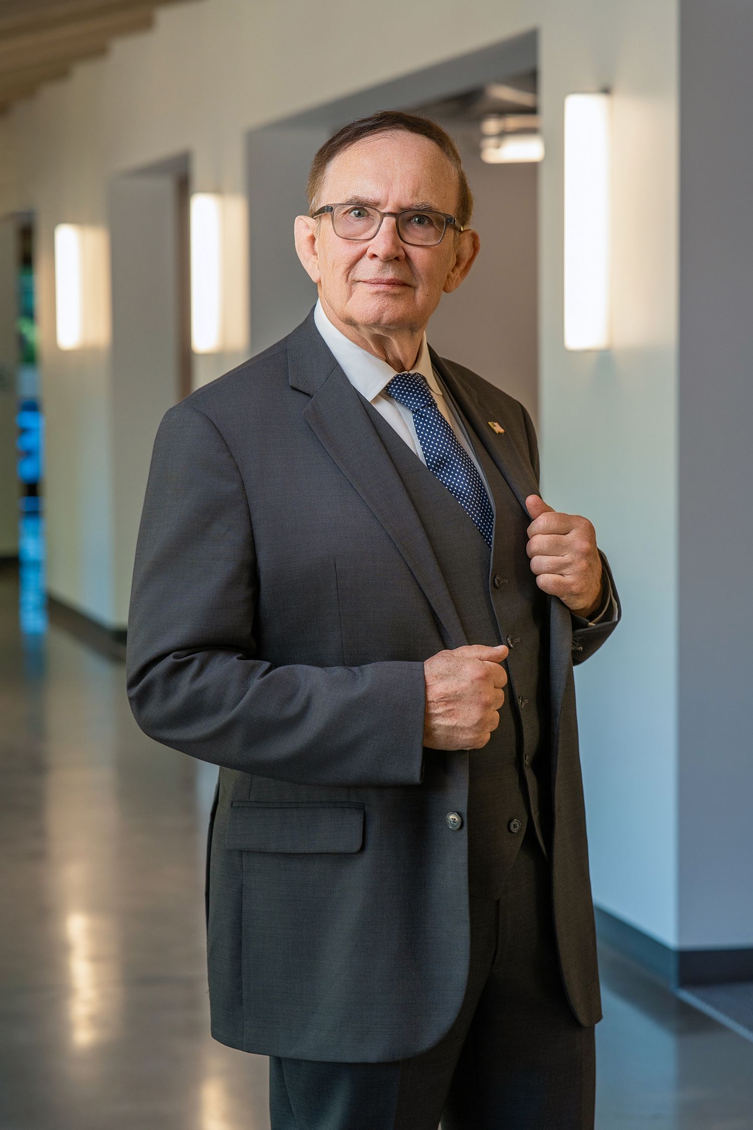 Elderly businessman in gray suit and tie poses confidently in modern hallway with glasses and lapel pin.