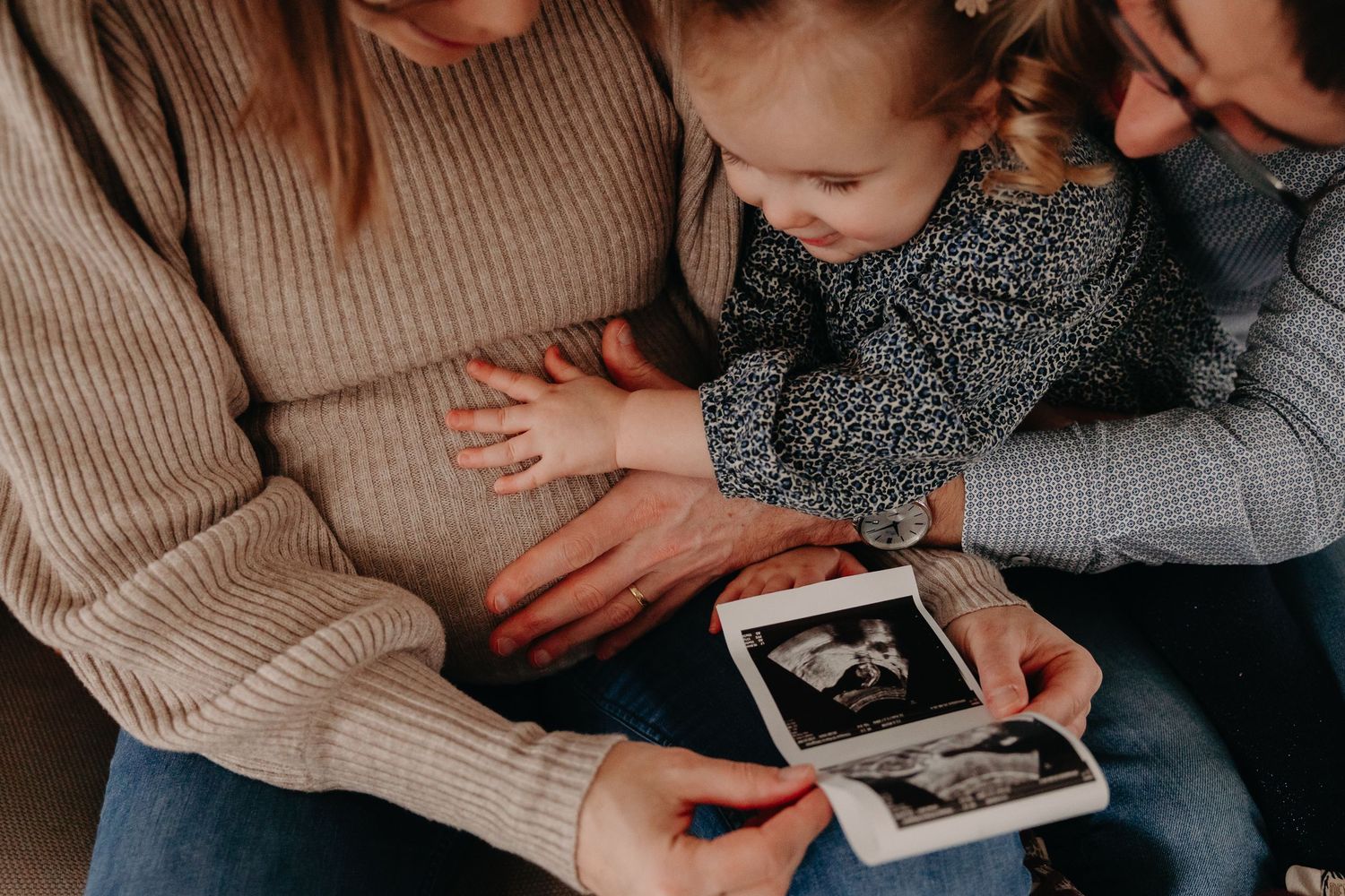 Family sitting together holding ultrasound photos, young toddler girl touching mother's pregnant belly.