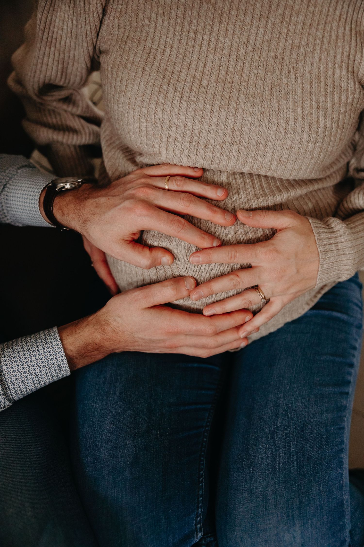 Couple tenderly holding hands together on pregnant belly, wearing rings and watch, maternity photo.