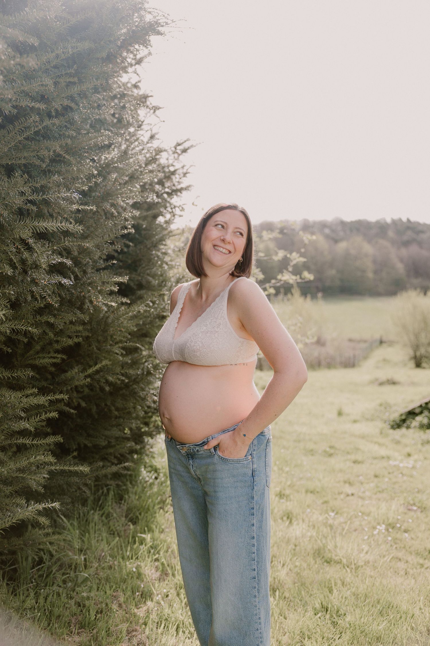 Smiling pregnant woman in beige bralette and jeans standing beside evergreen tree in sunny field.