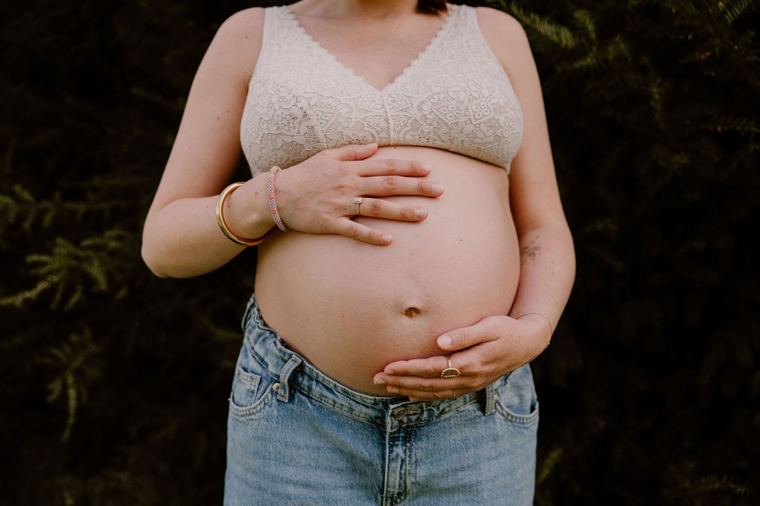 Close-up of pregnant woman in lace bralette and jeans holding bare baby bump with gold bracelet.