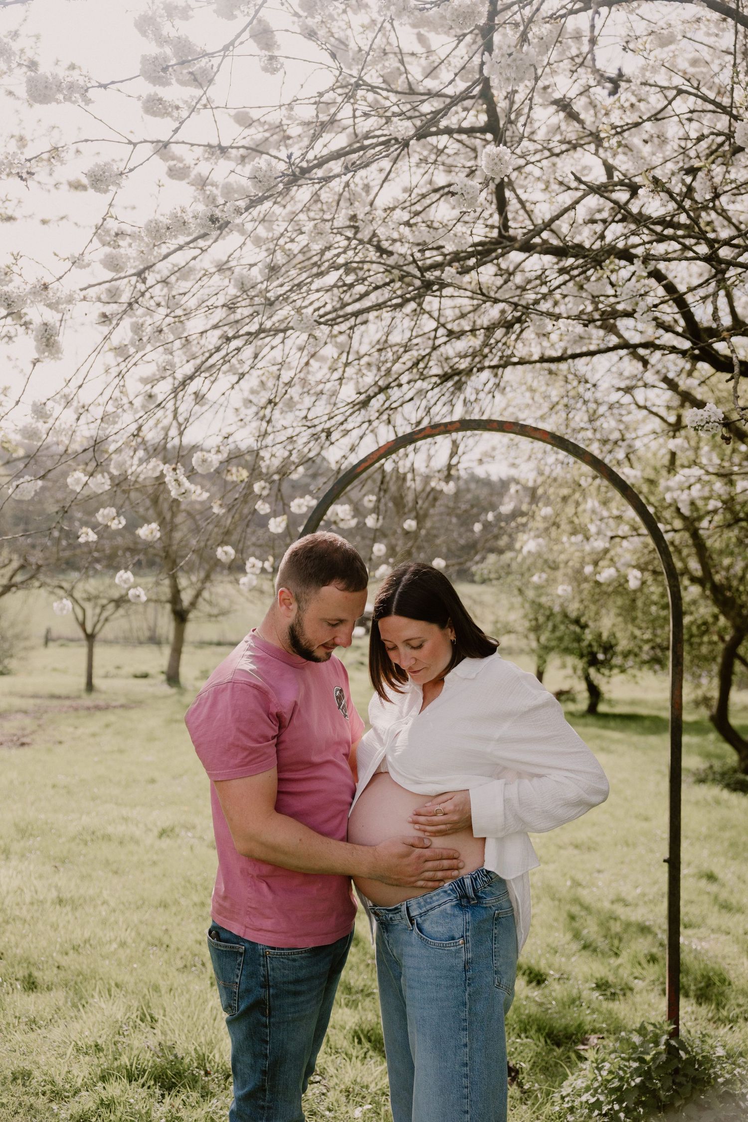 Couple standing under floral arch in blooming orchard, both looking down at pregnant woman's belly.