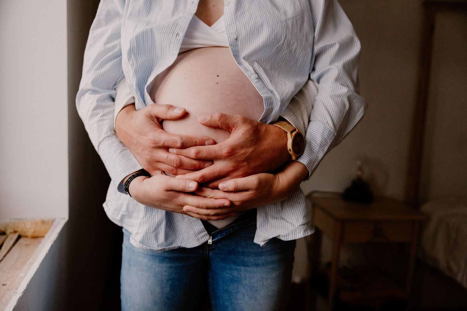 Close-up of couple's hands cradling pregnant belly, woman wearing open blue shirt and jeans.