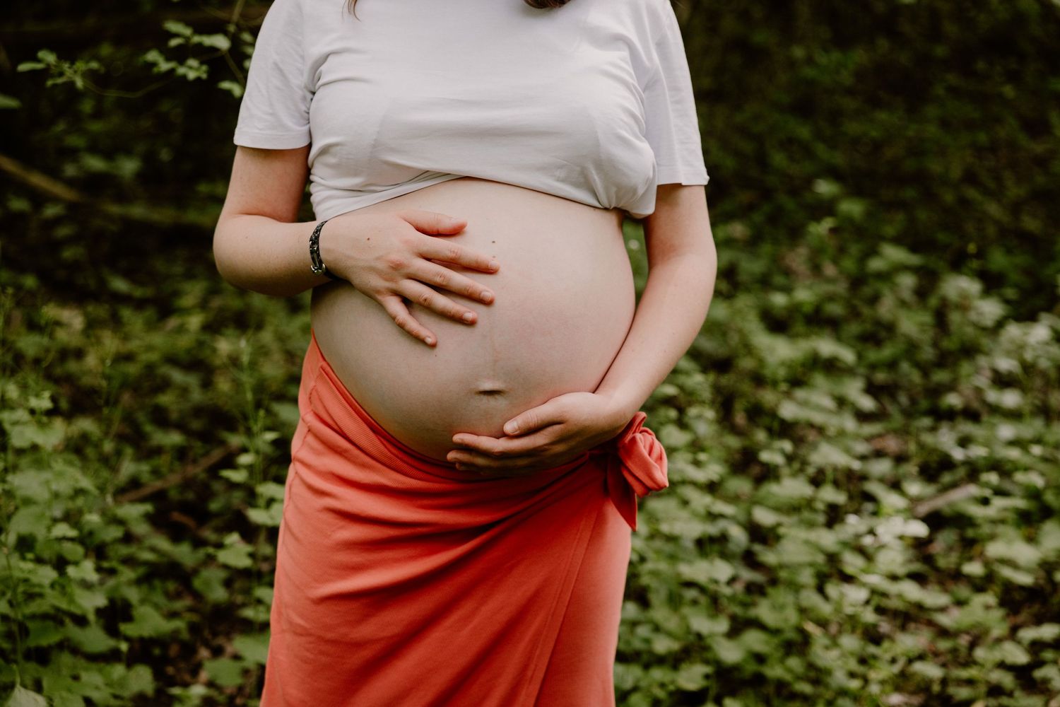 Close-up of pregnant woman in white top and coral skirt cradling bare baby bump outdoors.