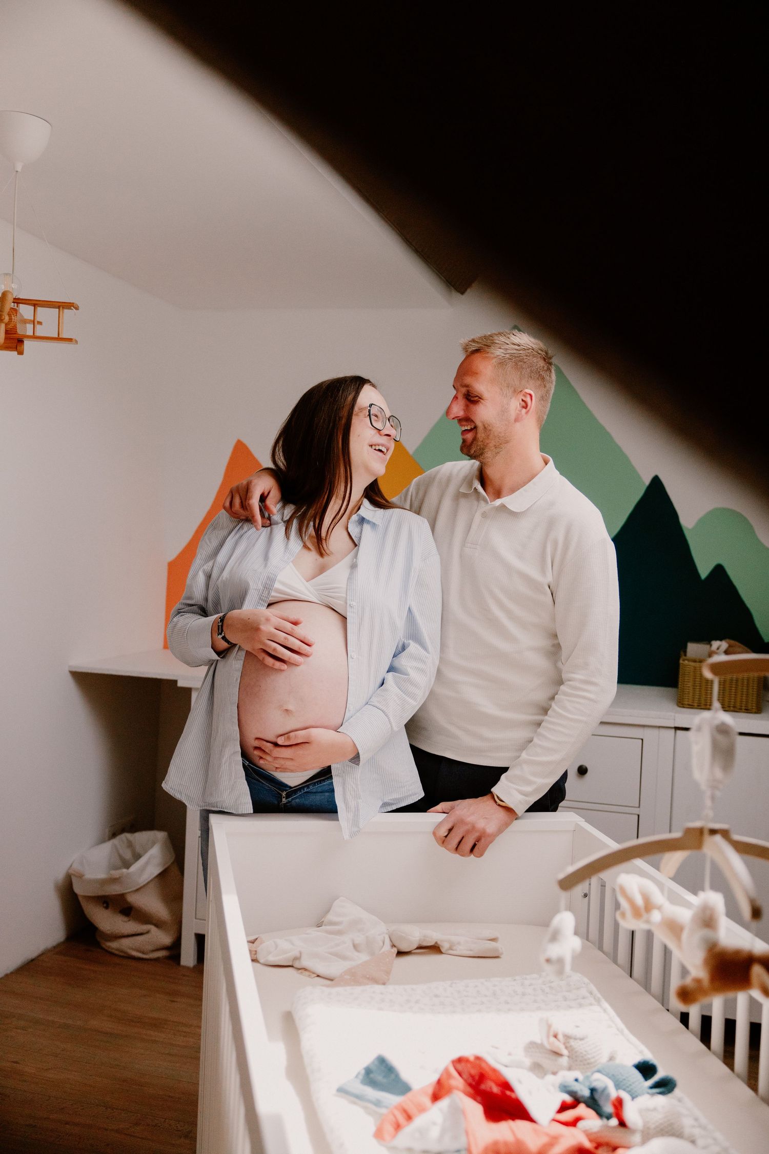 Happy couple in nursery room, pregnant woman holding baby bump while partner embraces her near decorated crib.