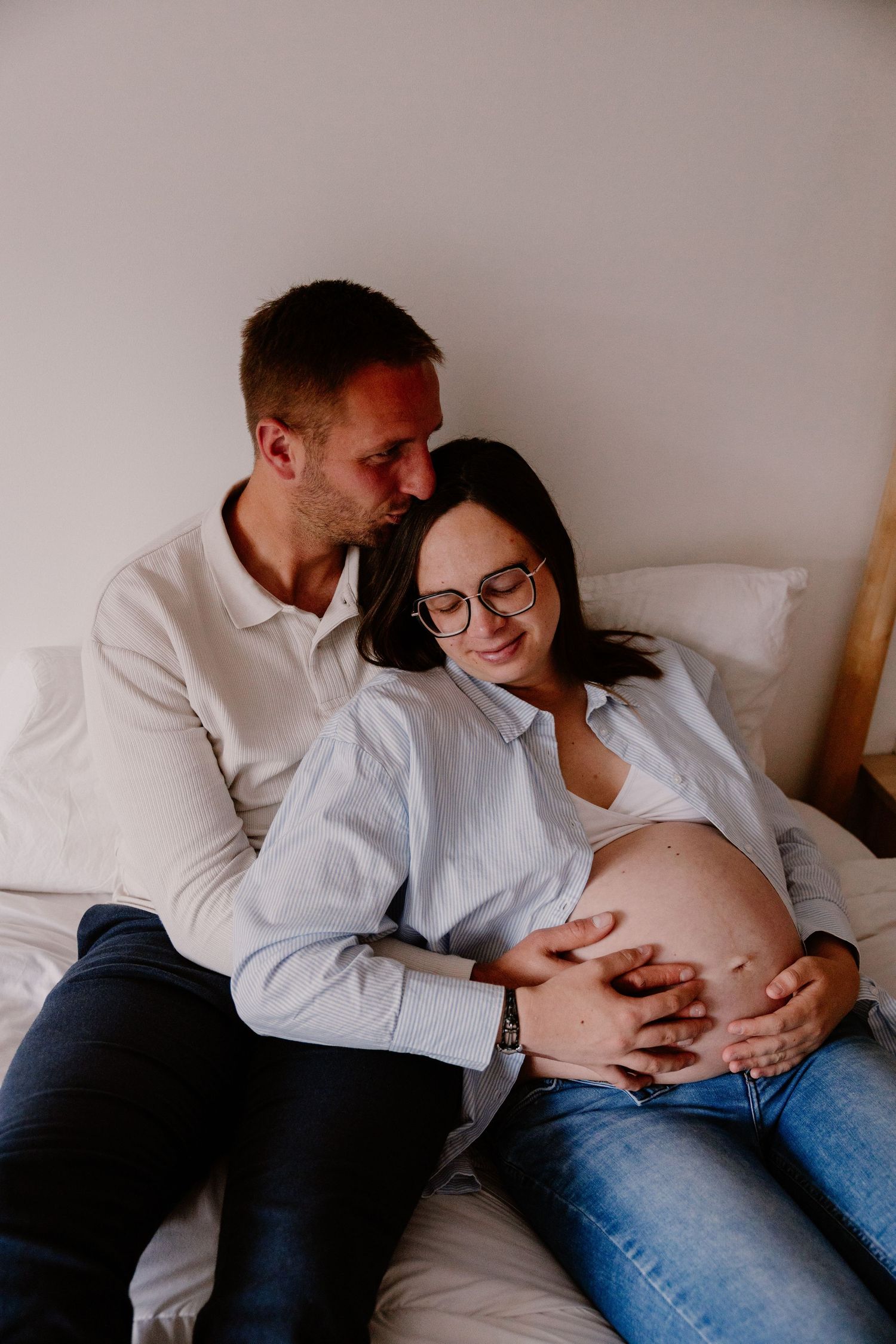 Couple sitting on bed, man kissing pregnant woman's head while both cradle her baby bump.