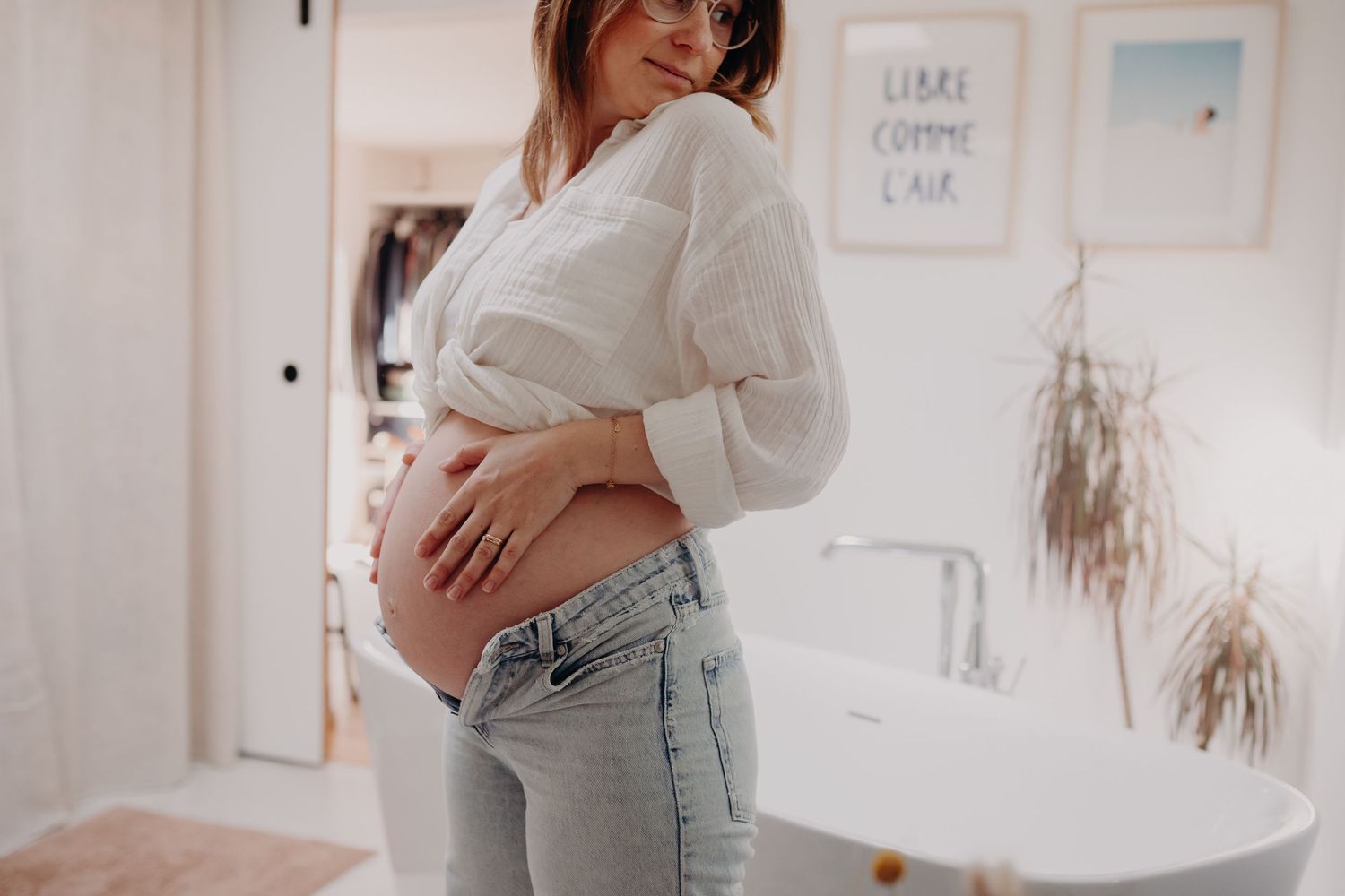 Smiling pregnant woman in white blouse and jeans cradling baby bump in bright bathroom.