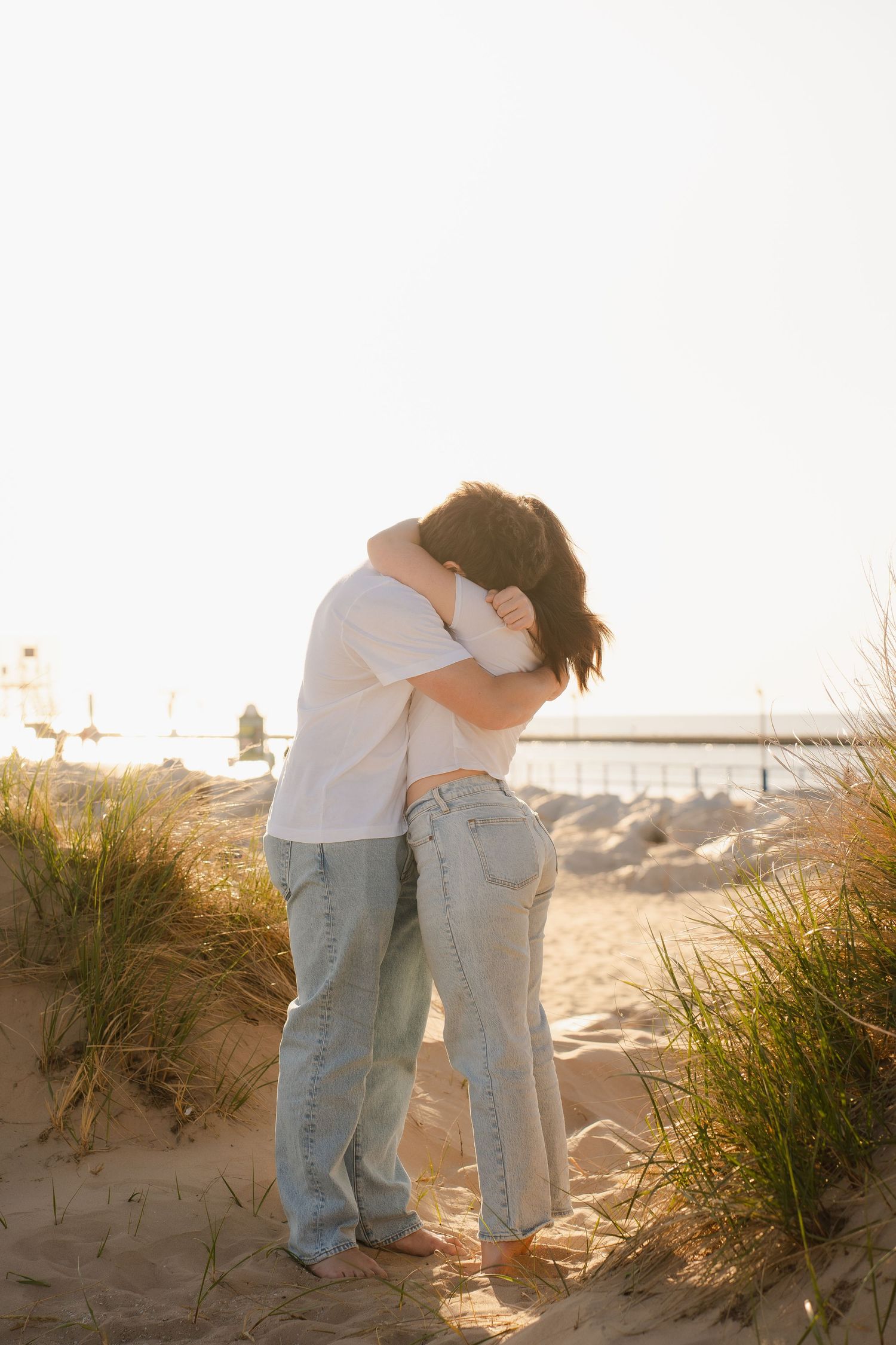 Couple embracing romantically on sandy beach dunes at golden hour, wearing jeans and white shirts.