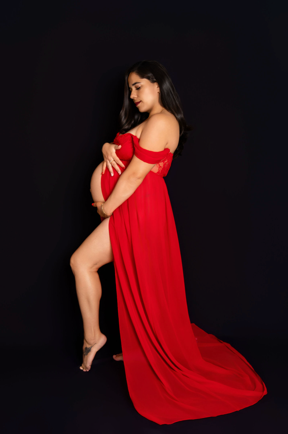 Pregnant woman in elegant red off-shoulder gown with train poses gracefully against black background.