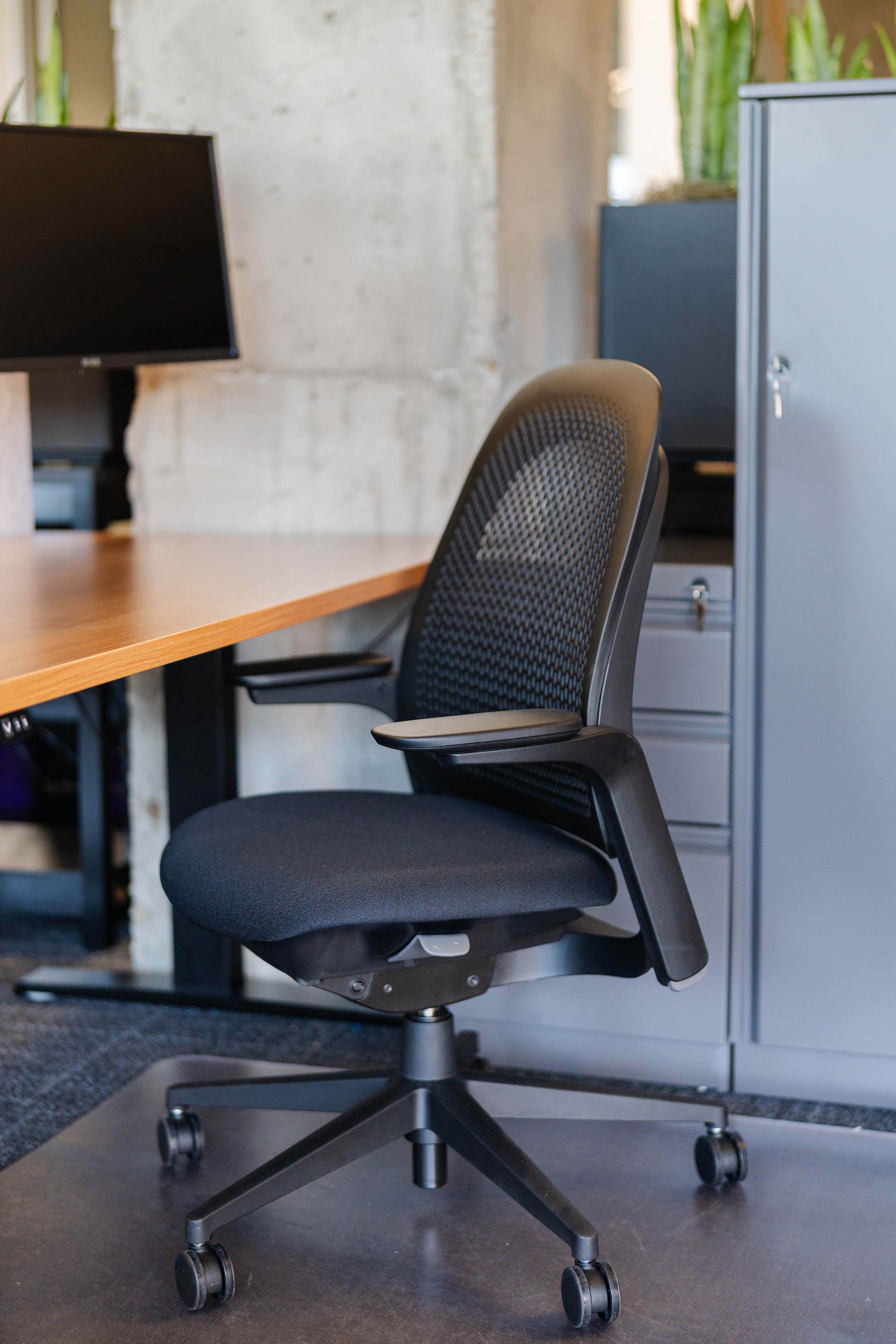 Black ergonomic mesh office chair with armrests on casters beside a wooden desk and gray filing cabinet.