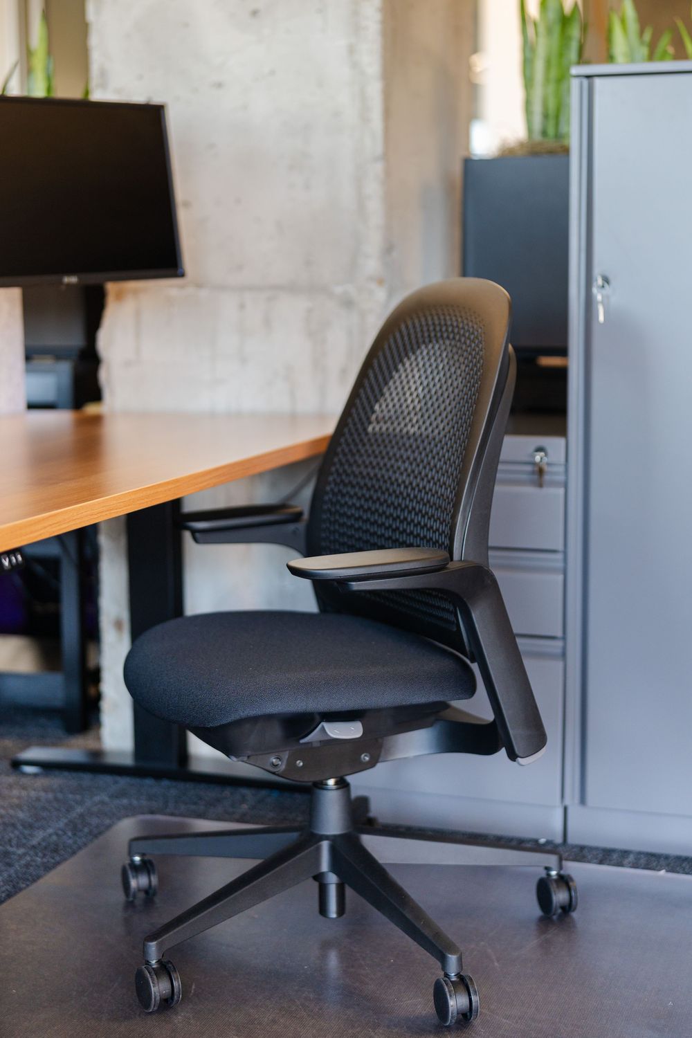 Black ergonomic mesh office chair with armrests on casters beside a wooden desk and gray filing cabinet.