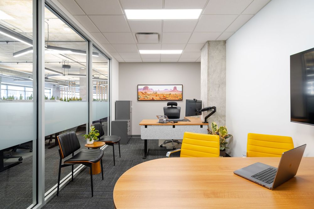 Modern private office with yellow chairs, standing desk, monitor, and glass partition walls in a contemporary workspace.