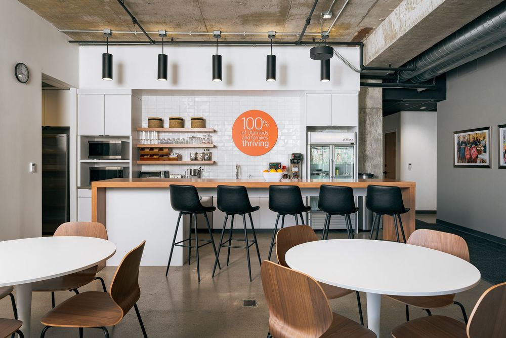 Stylish office kitchen and dining area with white cabinets, bar seating, pendant lights, and exposed concrete ceiling.