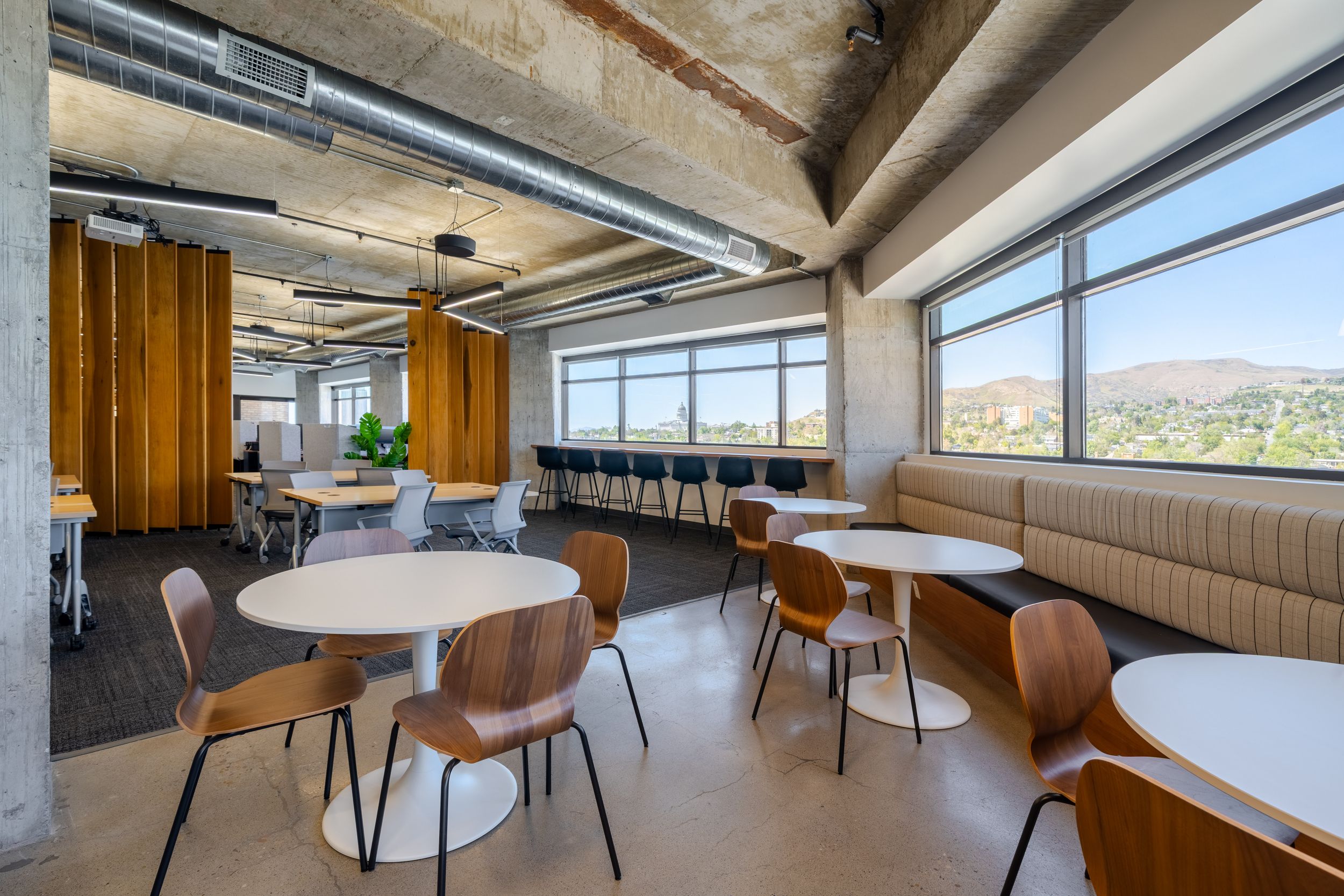 Modern office breakroom with round white tables, wooden chairs, booth seating, and large windows overlooking a scenic hillside view.