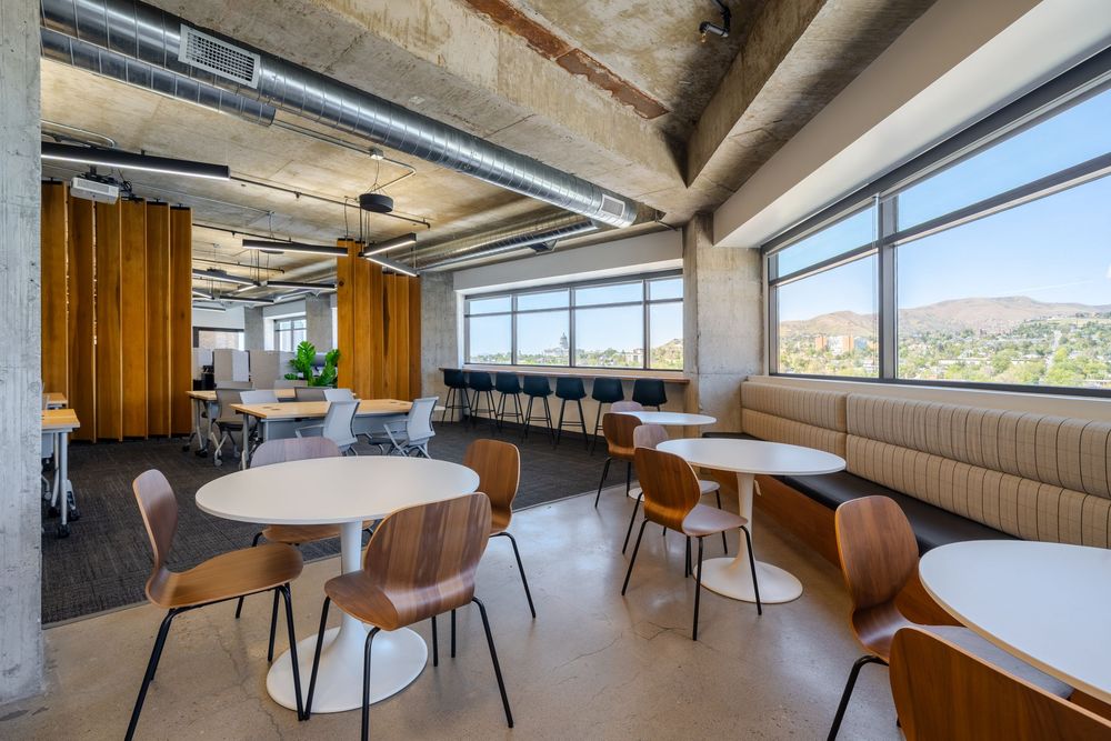 Modern office breakroom with round white tables, wooden chairs, booth seating, and large windows overlooking a scenic hillside view.