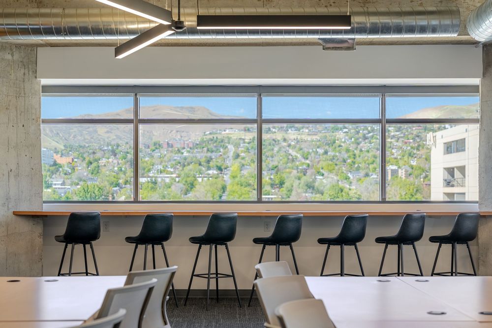Modern office space with black bar chairs at a wooden counter overlooking a panoramic city and hillside view.
