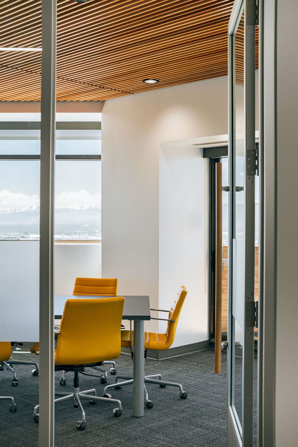 Modern conference room with yellow chairs, wood slat ceiling, glass walls, and mountain views.
