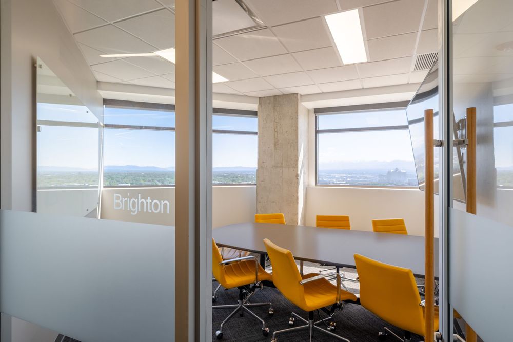 Modern conference room with yellow chairs, oval table, glass walls, and panoramic city views through large windows.