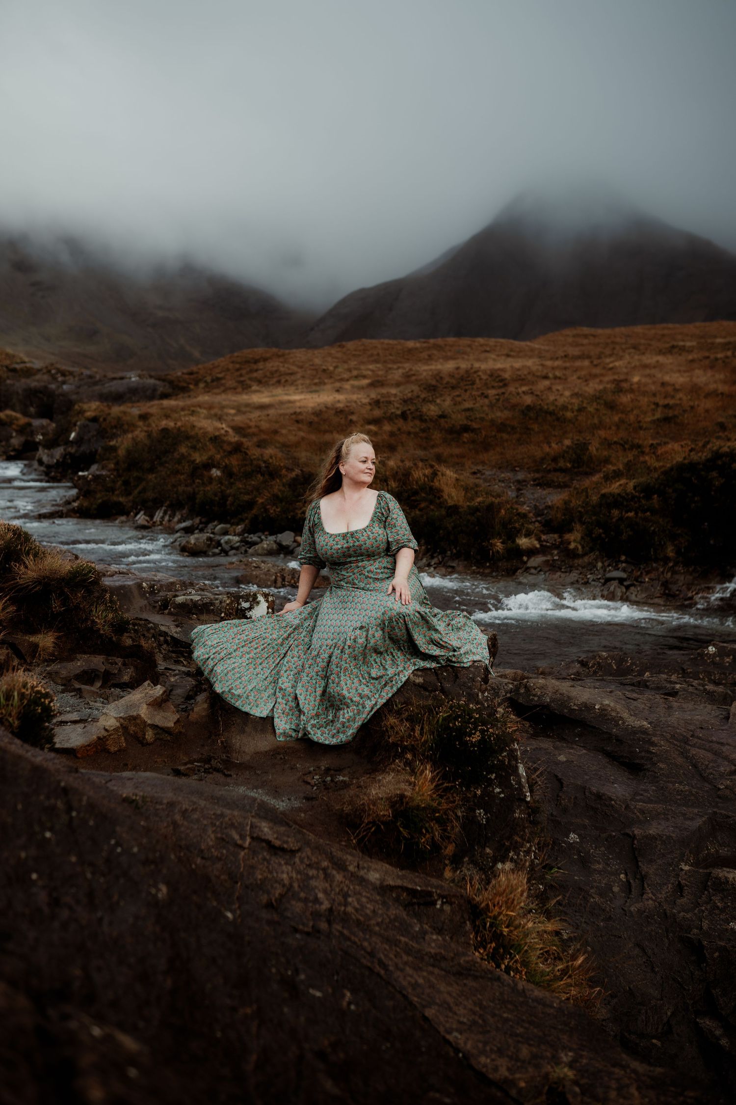 Woman in green dress sits on rocks by a misty Scottish highland stream with dramatic foggy mountains behind her.