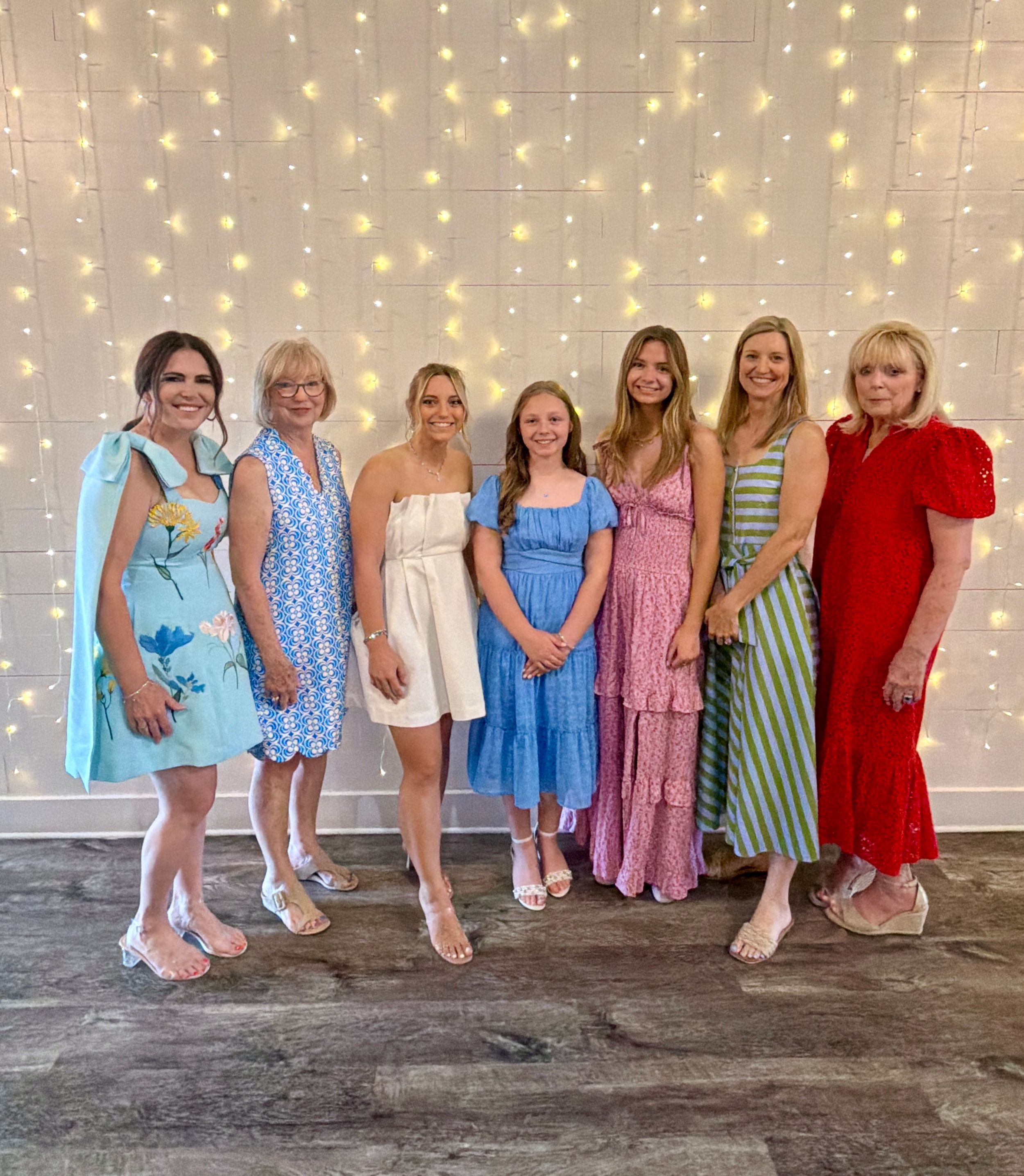 Seven women in colorful dresses pose together smiling in front of a twinkling fairy light backdrop.