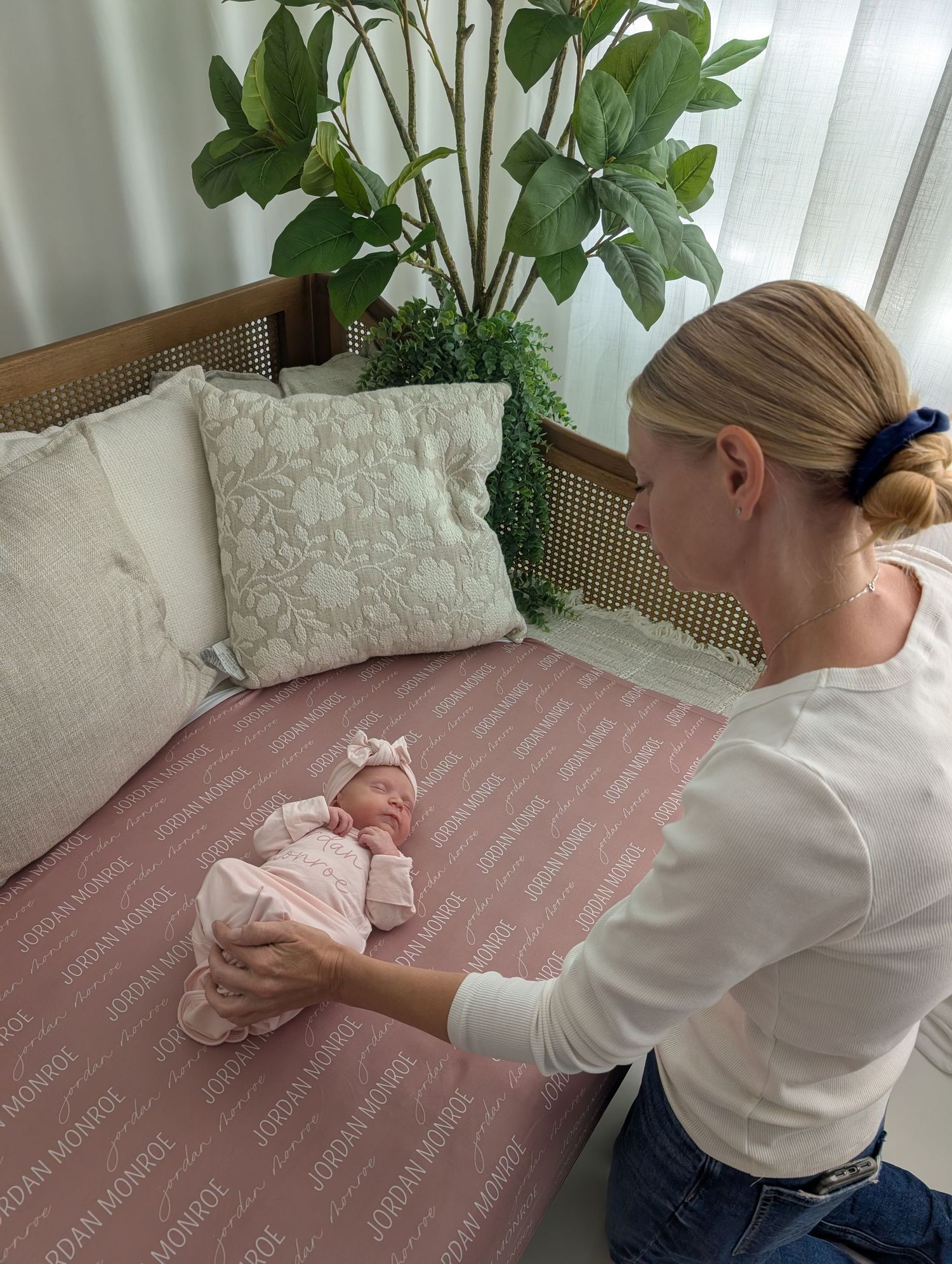 A woman in white top gently holds a newborn baby in pink outfit on a personalized pink blanket near a couch.