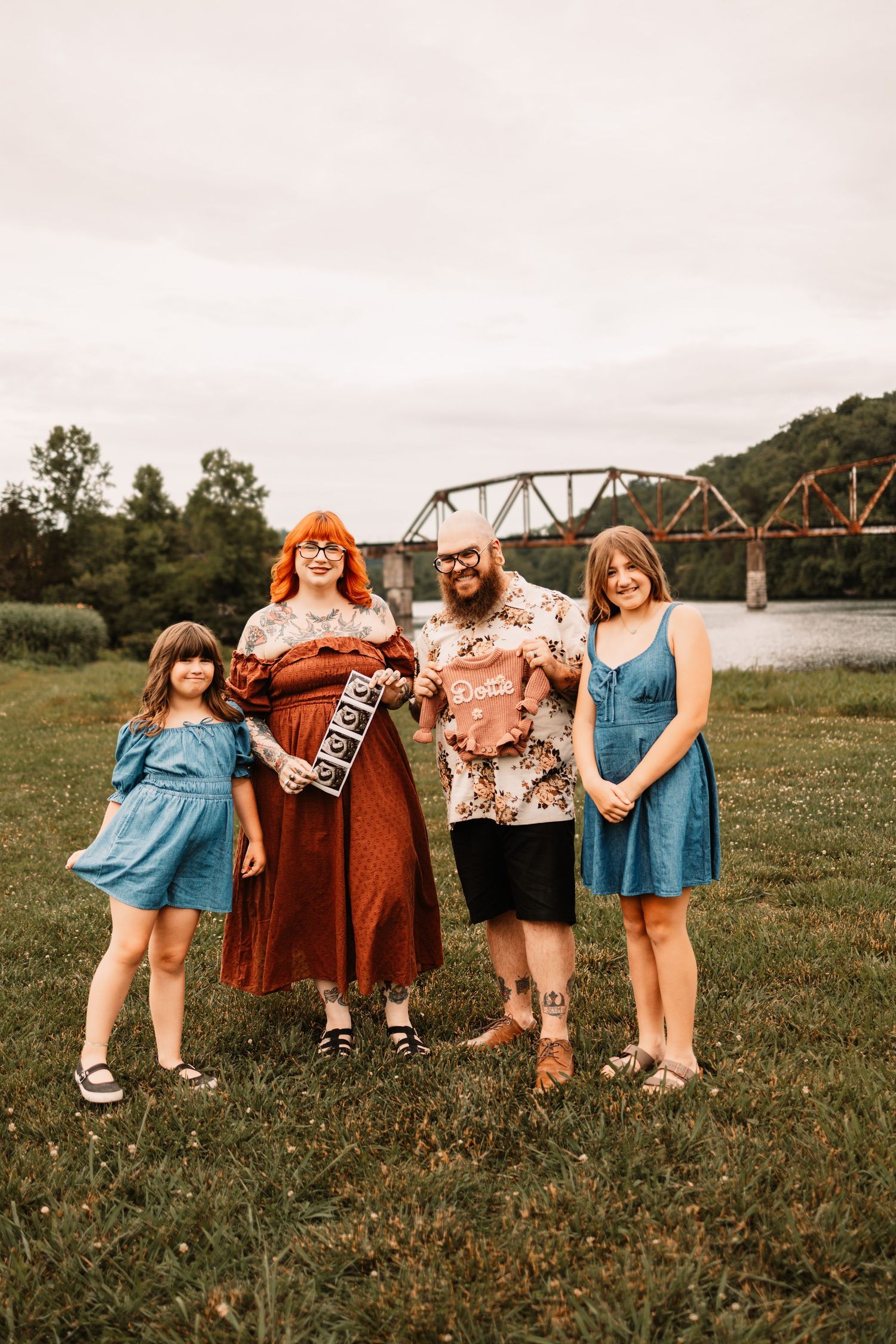Family of four smiling outdoors near a river bridge, holding ultrasound photos, announcing a pregnancy.