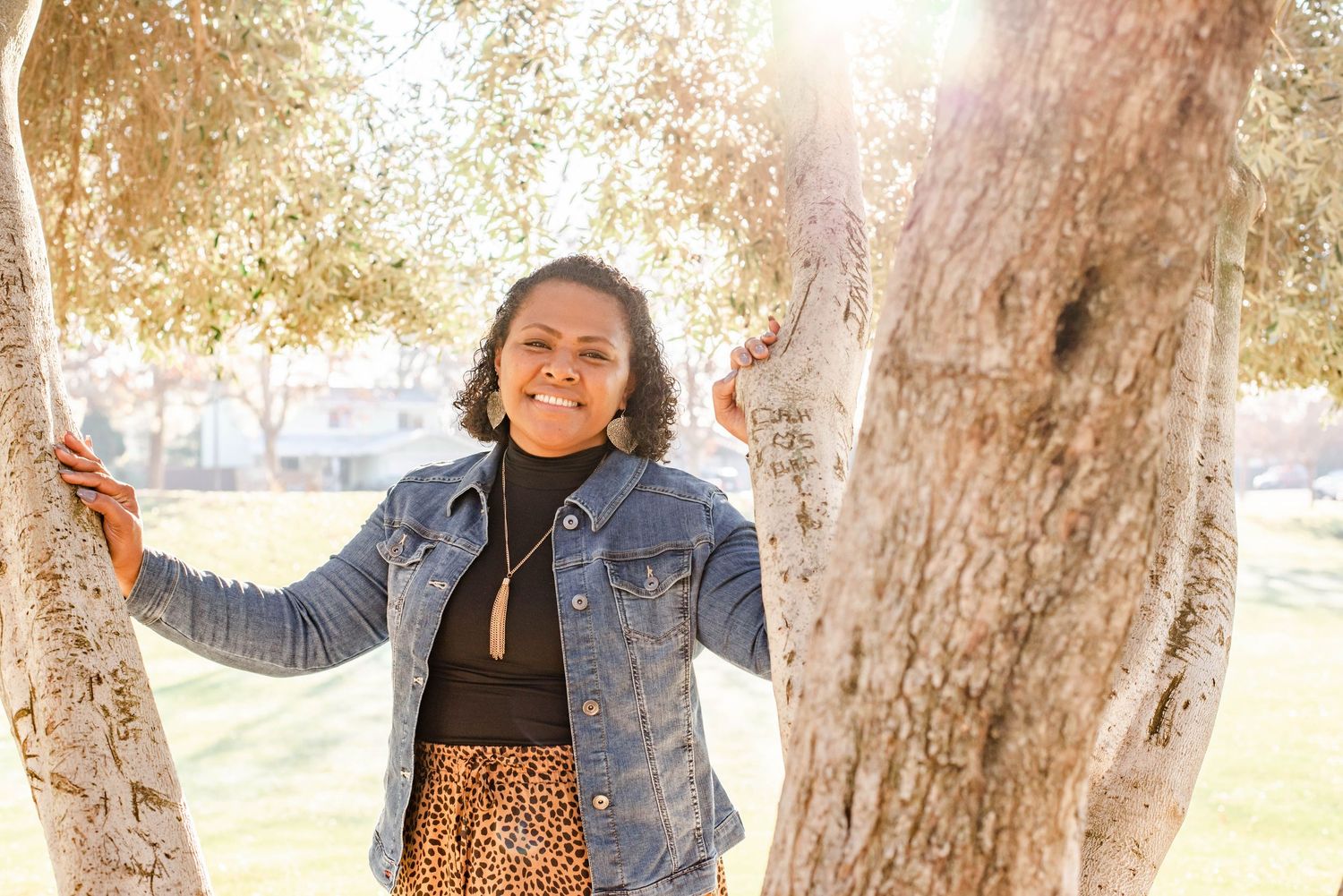 Young woman in denim jacket and leopard skirt smiling between trees in a sunny park.