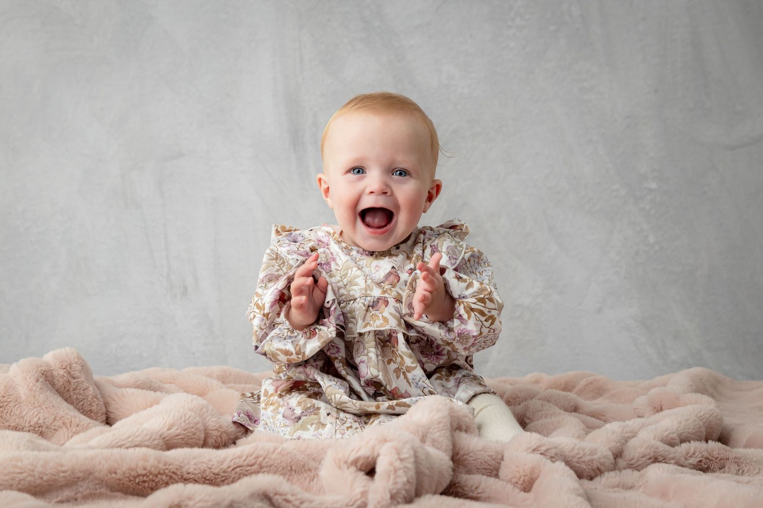 Happy baby in floral outfit sitting on pink fluffy blanket against gray wall, clapping hands and smiling.