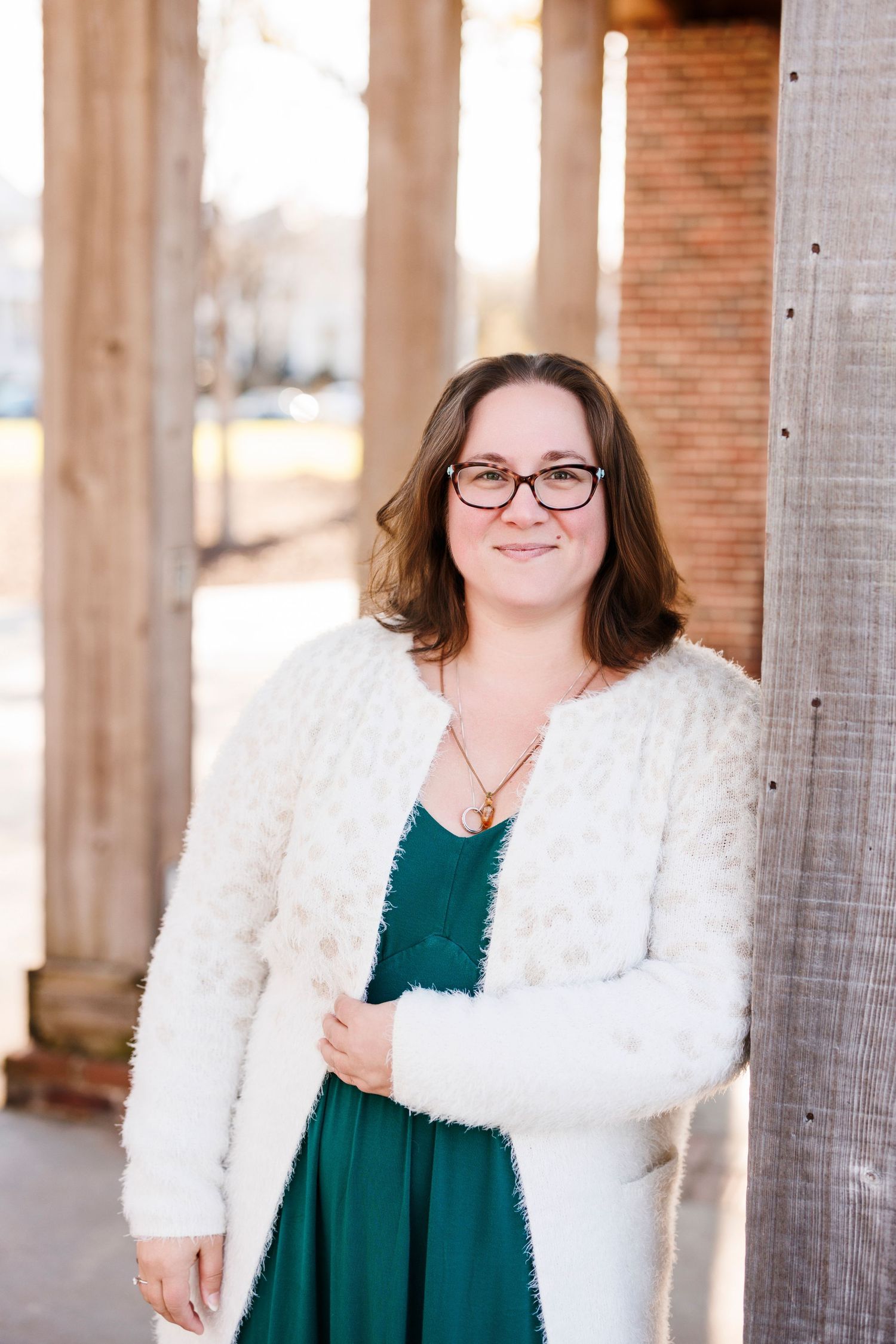 Woman in white fuzzy cardigan and green dress smiling outdoors near brick columns.
