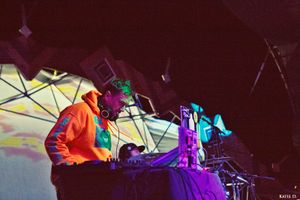 Close-up of DJ mixing at turntables wearing orange hoodie under psychedelic stage lighting.