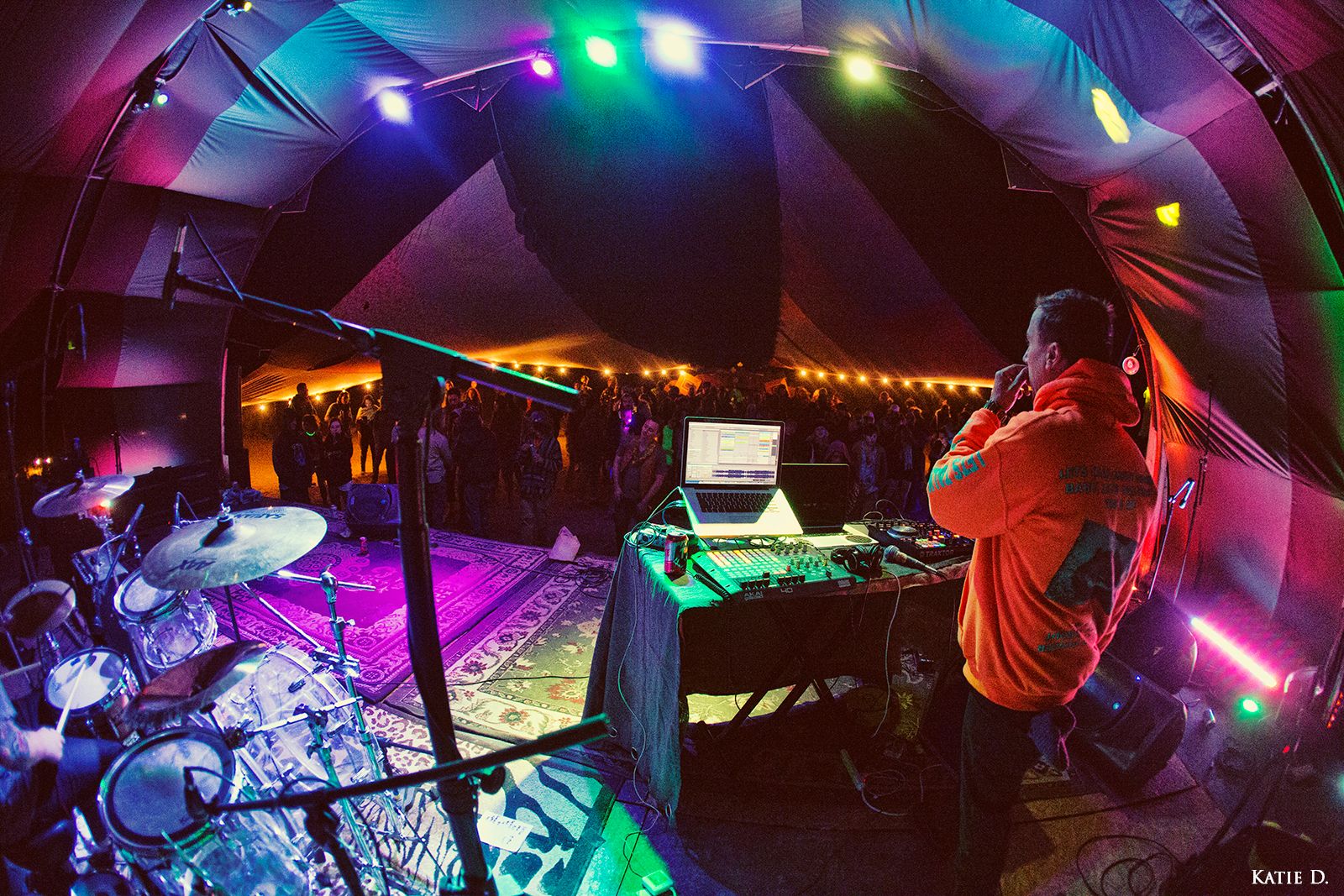 Wide-angle view of DJ and drummer performing on stage in colorful tent venue for live audience.