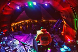DJ in orange hoodie performing for large crowd under multicolored lights in festival tent.