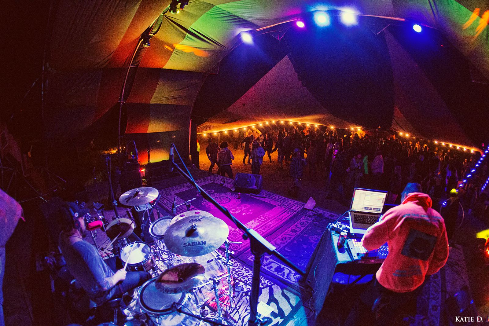 Overhead view of drummer and DJ performing under rainbow-colored tent lights for festival crowd.