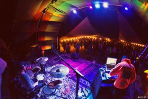 Overhead view of drummer and DJ performing under rainbow-colored tent lights for festival crowd.