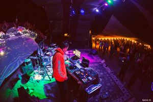 DJ in orange hoodie working mixing board on stage under blue and green festival lights.