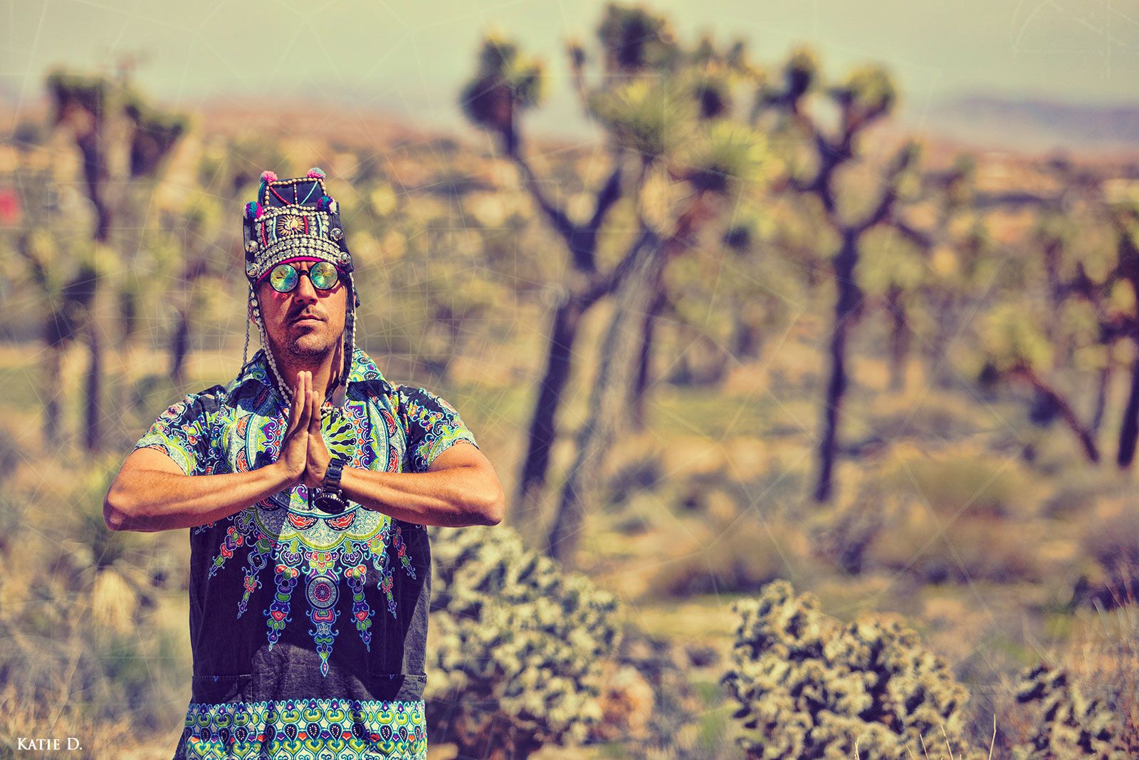 Man in colorful festival attire meditates with prayer hands in Joshua Tree desert.