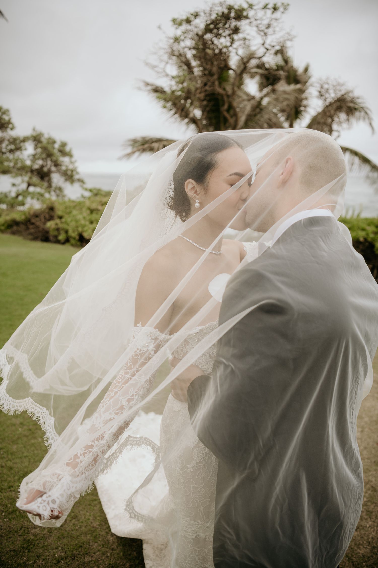 Bride and groom sharing a romantic kiss under a flowing lace wedding veil outdoors on their wedding day.