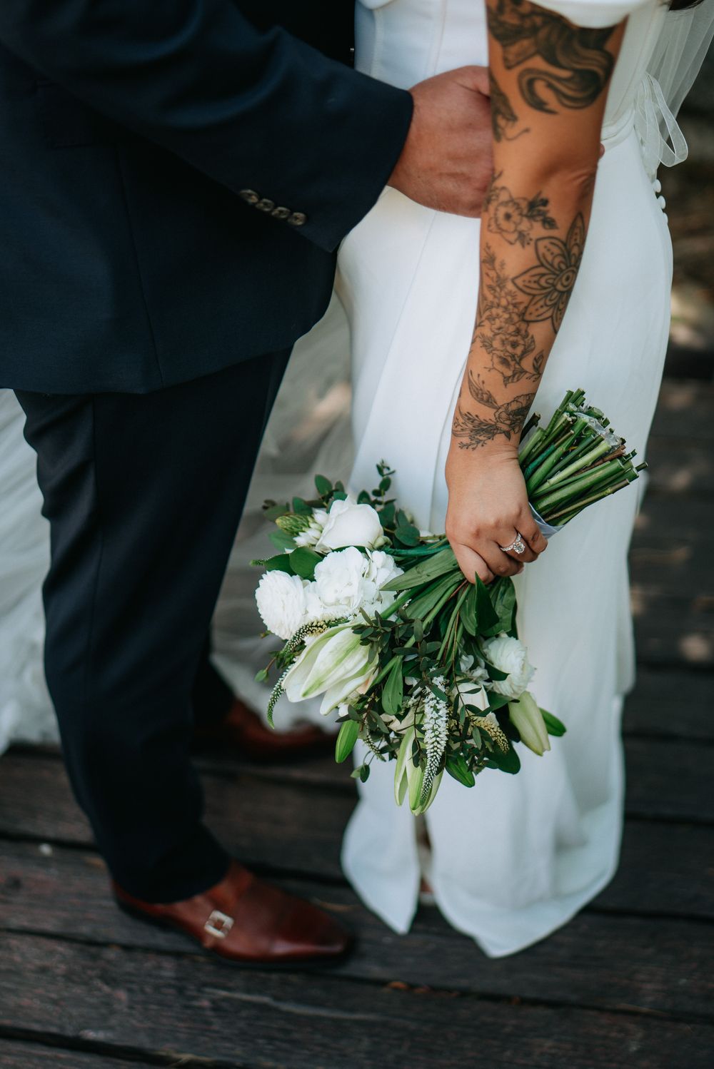 Bride with floral tattoo holds white bouquet of roses and tulips beside groom in navy suit on wooden deck.