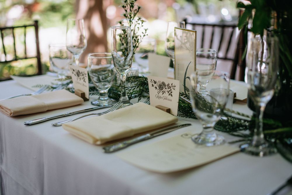 Elegant outdoor wedding table setting with white linens, greenery, crystal glassware, and decorative place cards.