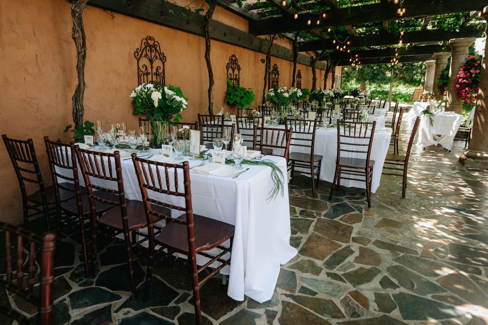 Elegant outdoor wedding reception with white-draped tables, greenery centerpieces, and dark Chiavari chairs on a stone patio.