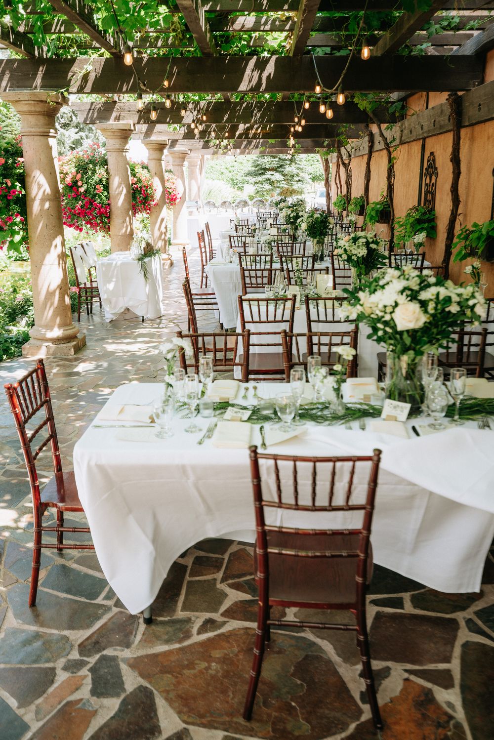 Elegant outdoor wedding reception with white tablecloths, chiavari chairs, floral centerpieces, and string lights under a pergola.