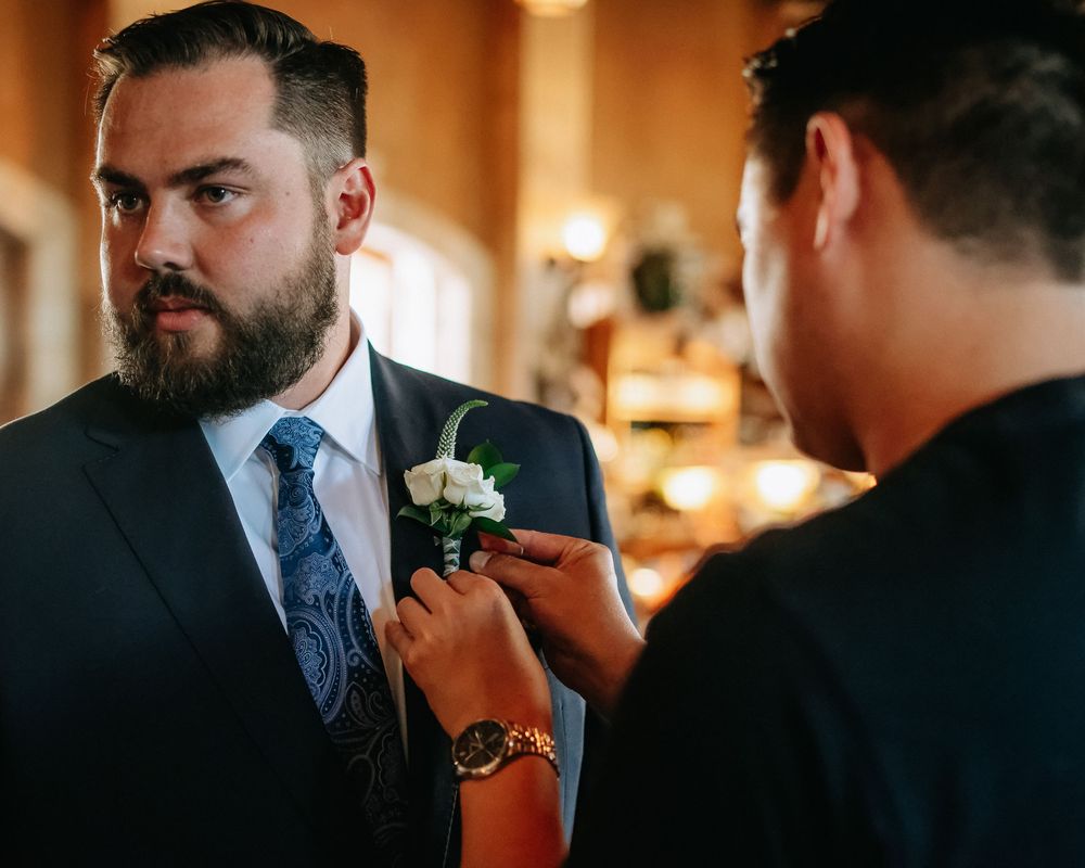 Groom in navy suit getting boutonniere pinned by groomsman before wedding ceremony.