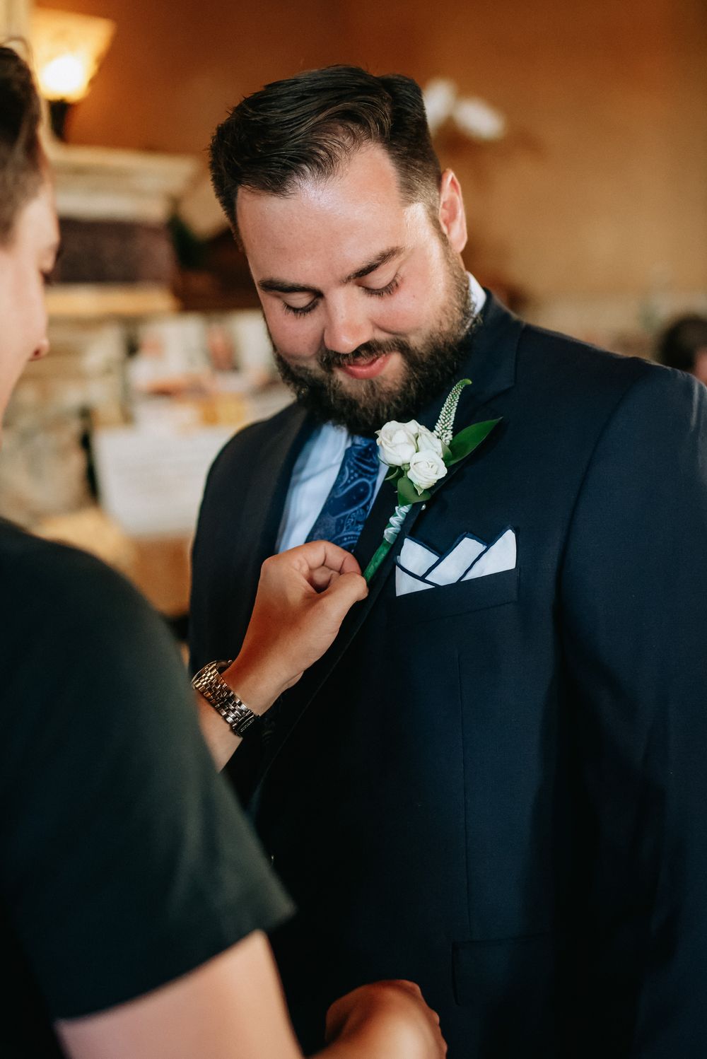 Groom in navy suit smiles as boutonniere with white roses is pinned to his lapel before wedding ceremony.