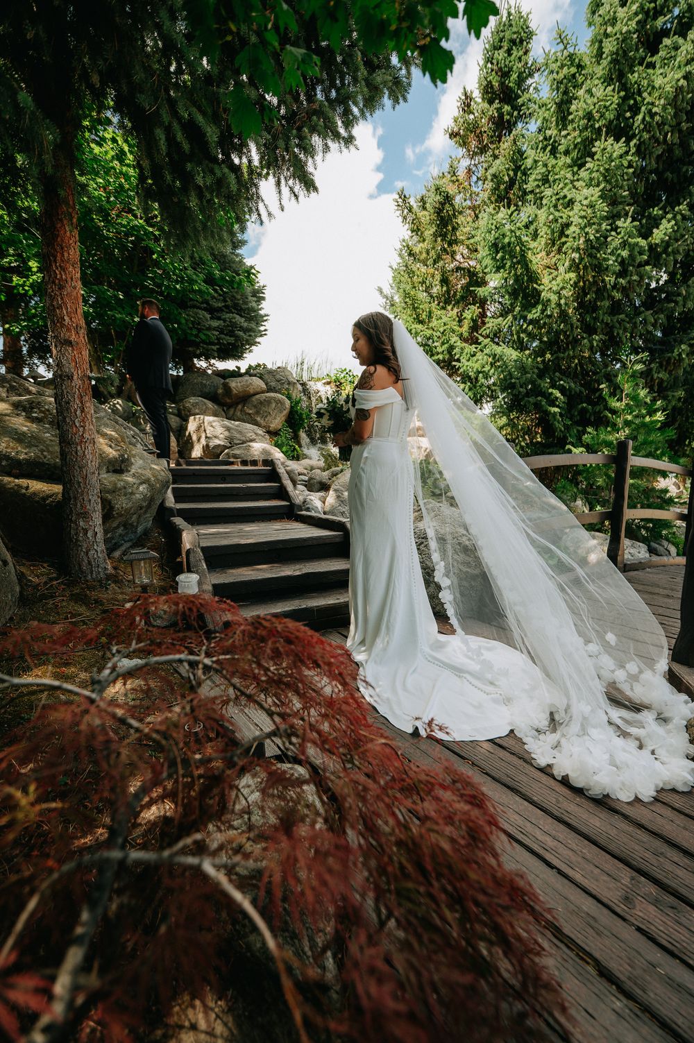 Bride in white off-shoulder gown with long veil stands on wooden deck beside stone steps in lush garden setting.