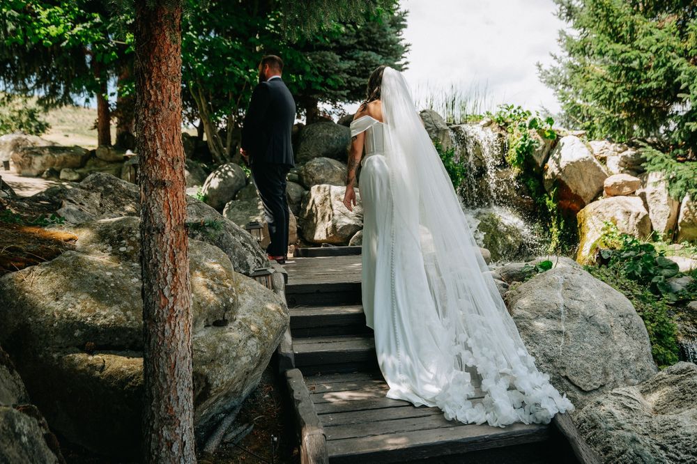 Bride in white gown and veil walks with groom up wooden steps surrounded by rocks and pine trees.